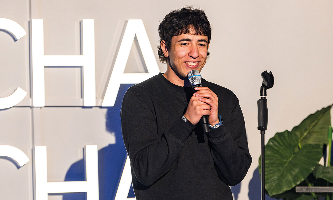 Ein junger Comedian steht auf einer Bühne und hält ein Mikrofon in der Hand. Er spricht lächelnd zum Publikum, während hinter ihm große Buchstaben einer Bühnenbeschriftung zu sehen sind. Ein Mikrofonständer steht neben ihm.