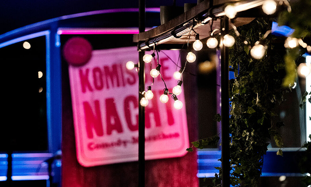 Ein beleuchtetes Schild mit der Aufschrift „Komische Nacht – Comedy-Marathon“ steht vor einer Bühne. Im Vordergrund hängen warme Lichterketten unter einem Pavillon, im Hintergrund leuchten bunte Bühnenlichter in Blau und Pink und schaffen eine abendliche Event-Atmosphäre.