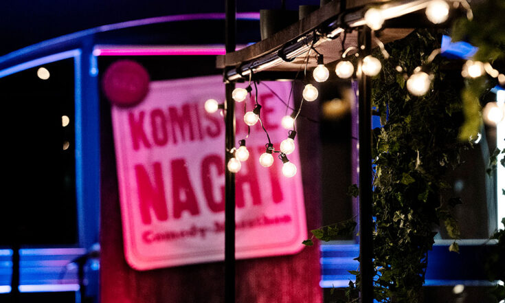 Ein beleuchtetes Schild mit der Aufschrift „Komische Nacht – Comedy-Marathon“ steht vor einer Bühne. Im Vordergrund hängen warme Lichterketten unter einem Pavillon, im Hintergrund leuchten bunte Bühnenlichter in Blau und Pink und schaffen eine abendliche Event-Atmosphäre.