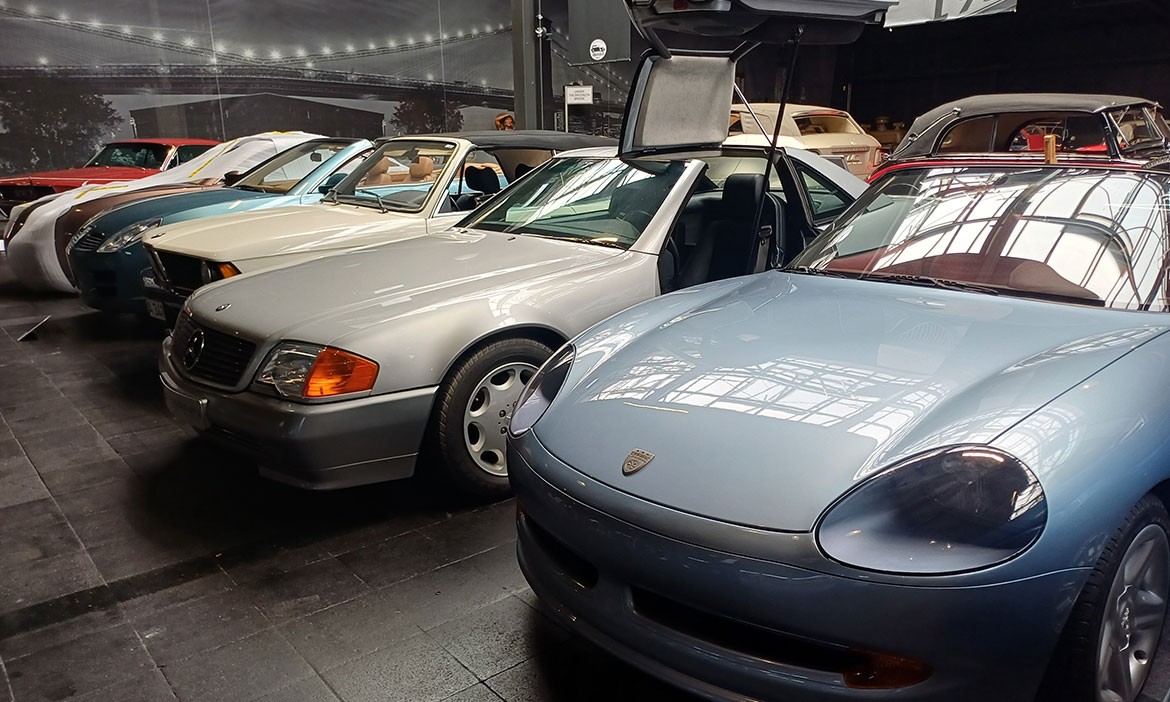 Mehrere Fahrzeuge in einer Ausstellungshalle des Automuseums Melle, darunter ein silberner Mercedes mit geöffneten Flügeltüren und ein hellblauer Porsche im Vordergrund. Im Hintergrund sind weitere Oldtimer und große Wandgrafiken zu sehen.