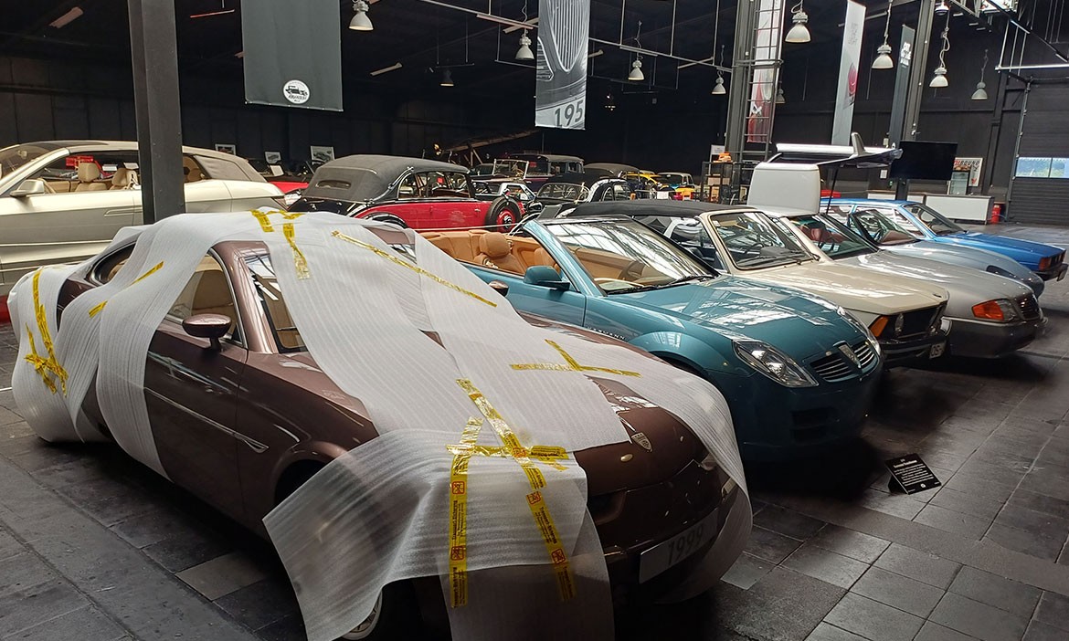 Blick in die Ausstellungshalle des Automuseums Melle mit mehreren Oldtimern und Cabriolets. Im Vordergrund steht ein teilweise mit Schutzfolie abgedecktes Fahrzeug, dahinter sind weitere historische Autos in Reihen zu sehen.