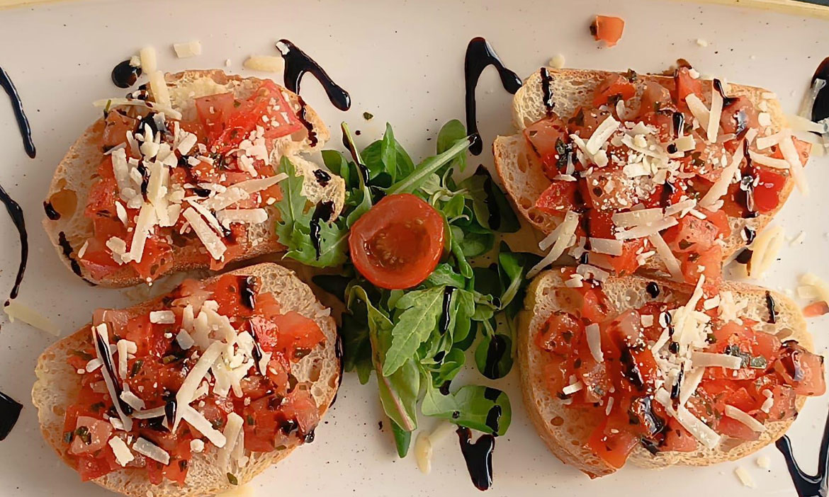 Vier Stück Bruschetta mit Tomatenwürfeln, gehobeltem Käse und dunkler Balsamico-Creme, angerichtet auf einem weißen Teller mit etwas Salat in der Mitte.