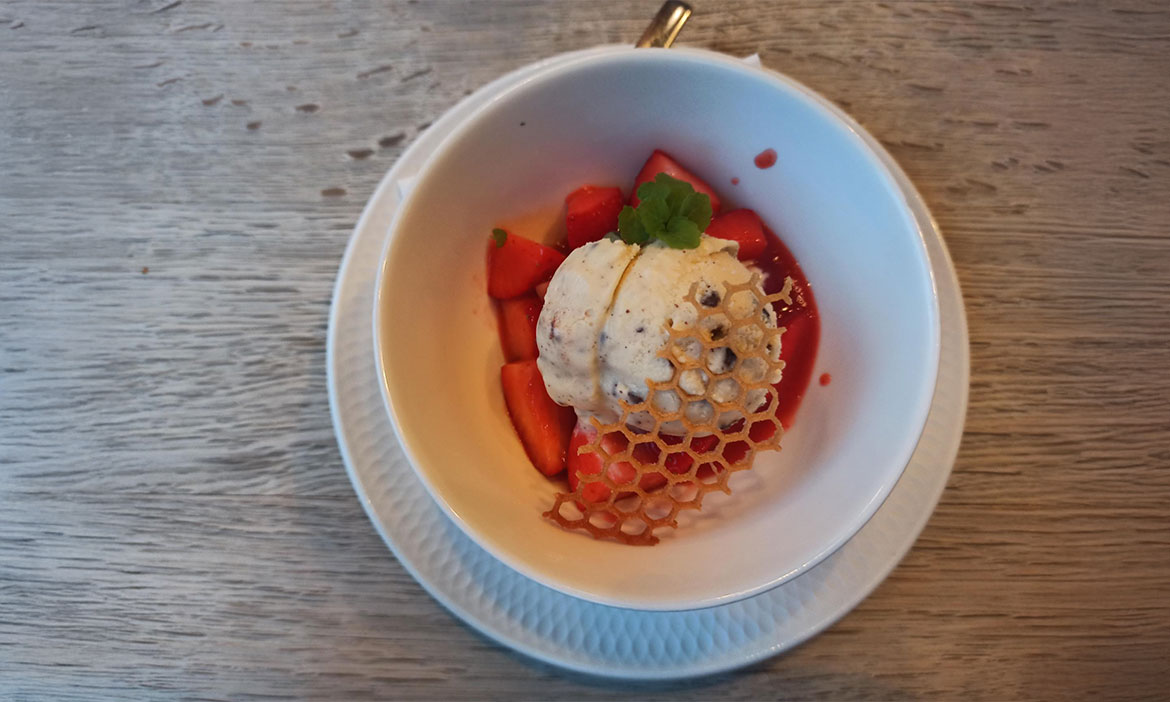 Dessert in einer weißen Schale: Erdbeerstücke mit einer Kugel Eis, garniert mit einer filigranen Wabe aus Karamell und einem kleinen Minzblatt.