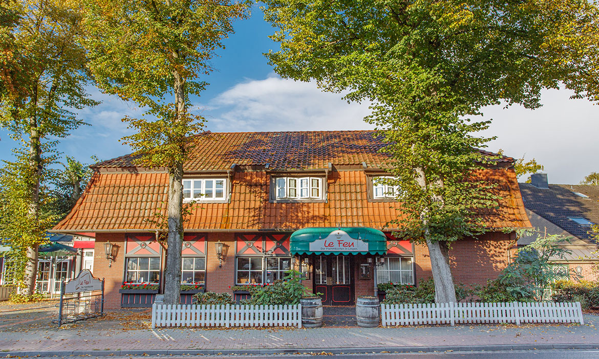 Außenansicht des Restaurants Le Feu in Lingen: ein backsteinfarbenes Haus mit rotem Ziegeldach, grüner Markise über dem Eingang und hohen Bäumen davor.