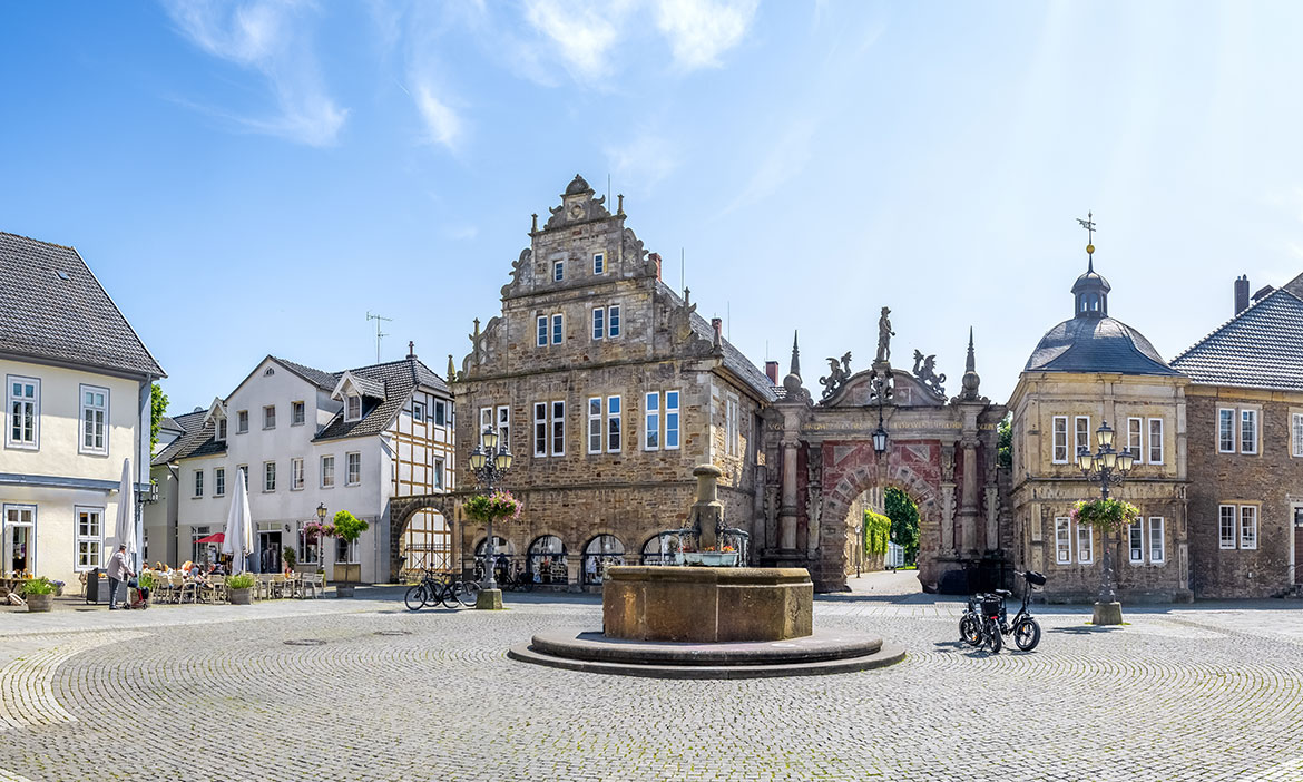 Historischer Marktplatz mit hellem Kopfsteinpflaster und rundem Brunnen in der Mitte. Im Hintergrund steht ein repräsentatives steinernes Gebäude mit geschwungenem Giebel, daneben ein reich verziertes Tor mit Figuren und rechts ein rundes Eckgebäude mit Kuppeldach. Links und rechts säumen helle Wohn- und Geschäftshäuser den Platz, vereinzelt sind Fahrräder, Straßenlaternen, Blumenkübel und sitzende Menschen zu sehen. Darüber ein blauer Himmel mit feinen Wolken.