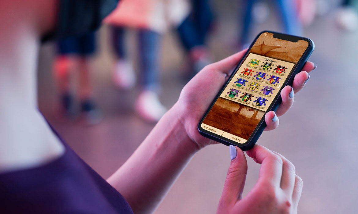 Nahaufnahme einer Person, die ein Smartphone mit beiden Händen hält. Auf dem Display ist eine App mit historisch gestalteter Oberfläche zu sehen; dargestellt sind mehrere farbige Wappen in einer Rasteransicht. Unten im Bildschirm stehen die Menüpunkte „Aufgaben“ und „Karte“. Der Hintergrund ist stark unscharf und zeigt mehrere Personen im Freien.