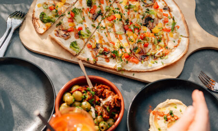 Auf einem gedeckten Tisch liegen ein großer Flammkuchen mit buntem Gemüse, Schälchen mit Oliven und Dip sowie schwarze Teller im Sonnenlicht.