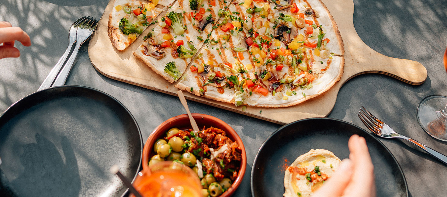 Auf einem gedeckten Tisch liegen ein großer Flammkuchen mit buntem Gemüse, Schälchen mit Oliven und Dip sowie schwarze Teller im Sonnenlicht.