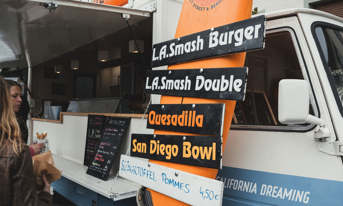 Ein Foodtruck mit kalifornischem Streetfood steht auf einem Festivalgelände. Vor dem geöffneten Verkaufsfenster hängen schwarze Menüschilder mit Angeboten wie „L.A. Smash Burger“, „Quesadilla“ und „San Diego Bowl“. Links wartet eine Besucherin mit Essenstüte vor dem Truck.