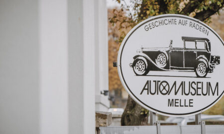 undes Schild des Automuseums Melle mit Oldtimer-Illustration und Schriftzug „Geschichte auf Rädern“ vor einem Eingangstor. Im Hintergrund sind Bäume und ein helles Gebäude unscharf zu sehen.