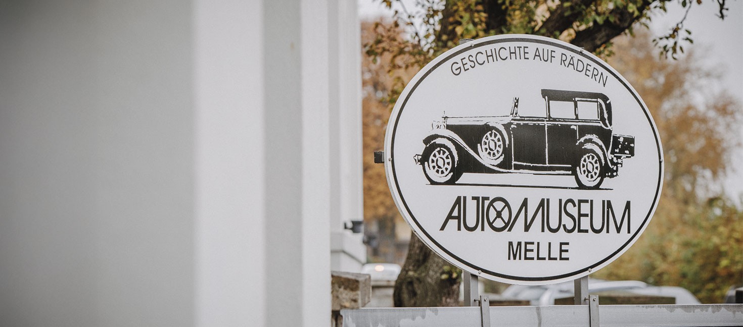 undes Schild des Automuseums Melle mit Oldtimer-Illustration und Schriftzug „Geschichte auf Rädern“ vor einem Eingangstor. Im Hintergrund sind Bäume und ein helles Gebäude unscharf zu sehen.