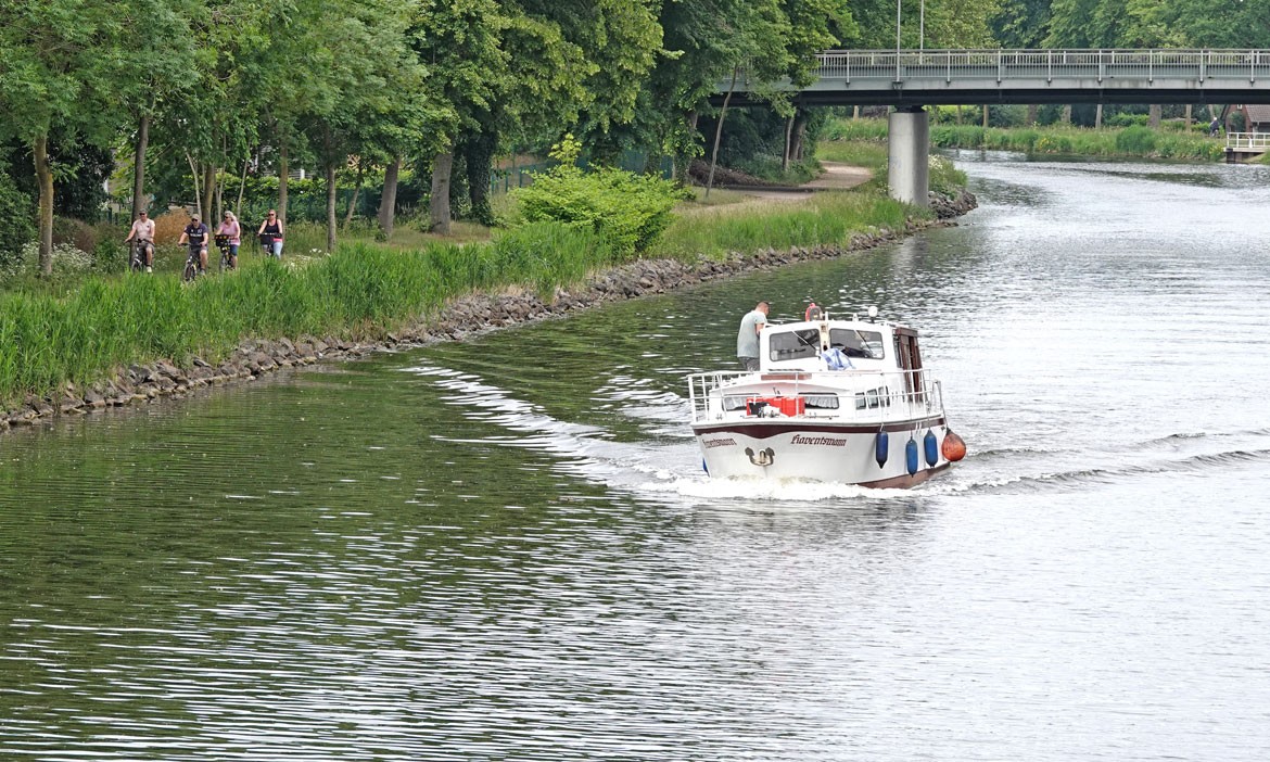 Ein kleines Motorboot fährt auf dem Dortmund-Ems-Kanal. Entlang des begrünten Ufers verläuft ein Radweg, auf dem mehrere Radfahrende unterwegs sind. Im Hintergrund überspannt eine Brücke den Kanal.