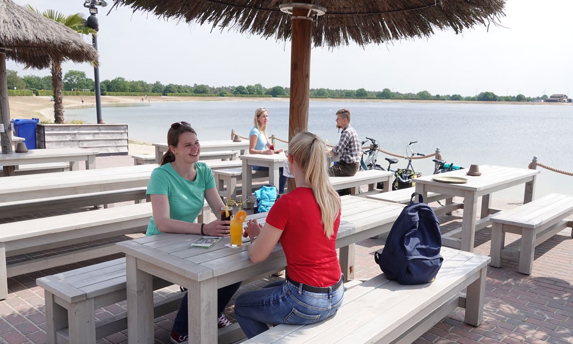 Menschen sitzen an hellen Holztischen auf einer Terrasse direkt am Ufer eines großen Sees. Im Hintergrund sind ein Sandstrand, Fahrräder und die weite Wasserfläche des Speichersees zu erkennen.
