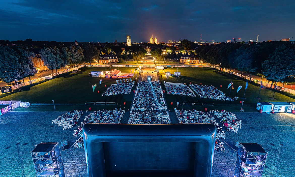 Ein gut besuchtes Open-Air-Kino bei Abenddämmerung. Viele Menschen sitzen in Reihen vor einer großen Leinwand, auf der ein Film läuft. Die Veranstaltung findet auf einem Platz zwischen historischen Gebäuden statt, die warm beleuchtet sind. Rechts neben der Leinwand steht ein Verkaufswagen.