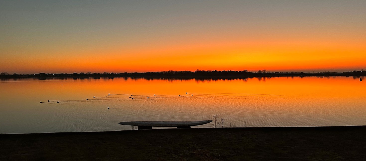 Stimmungsvoller Sonnenuntergang am Speichersee Geeste. Der Himmel leuchtet kräftig orange und spiegelt sich im ruhigen Wasser. Im Vordergrund sind eine Bank am Ufer und mehrere Wasservögel auf dem See zu sehen.