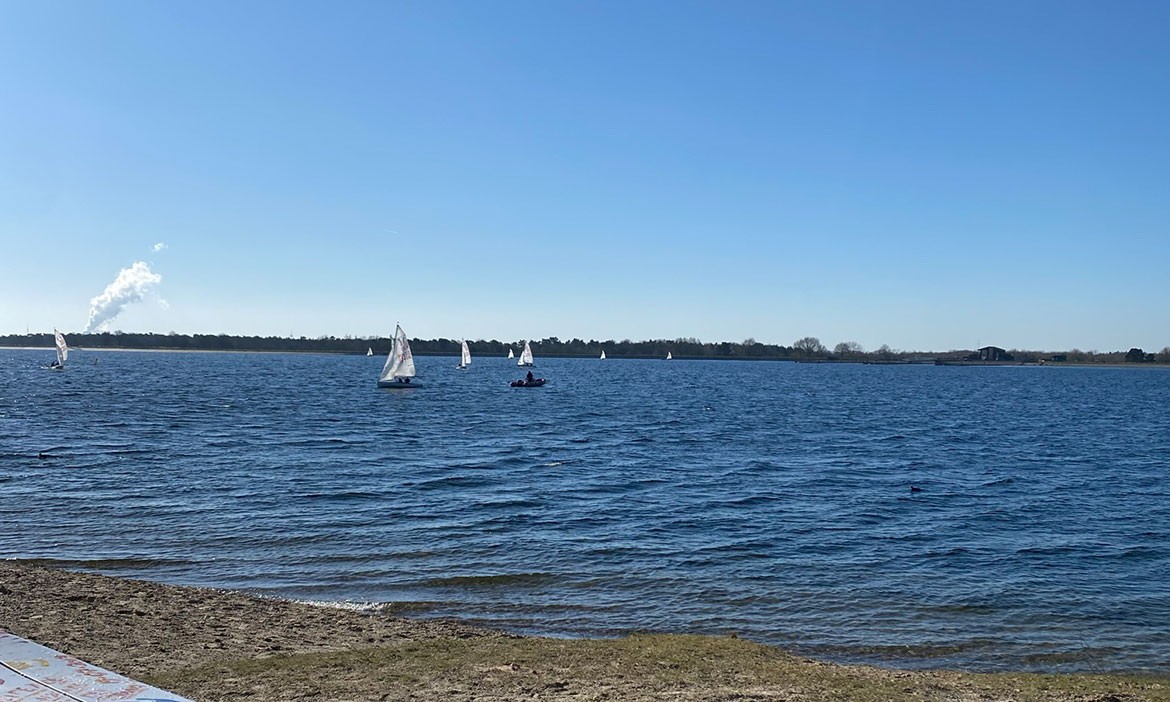 Weiter Blick über den Speichersee Geeste an einem klaren Tag. Auf dem leicht bewegten Wasser sind mehrere kleine Segelboote unterwegs. Im Vordergrund liegt ein schmaler Uferstreifen, am Horizont sind Bäume und einzelne Gebäude zu sehen.