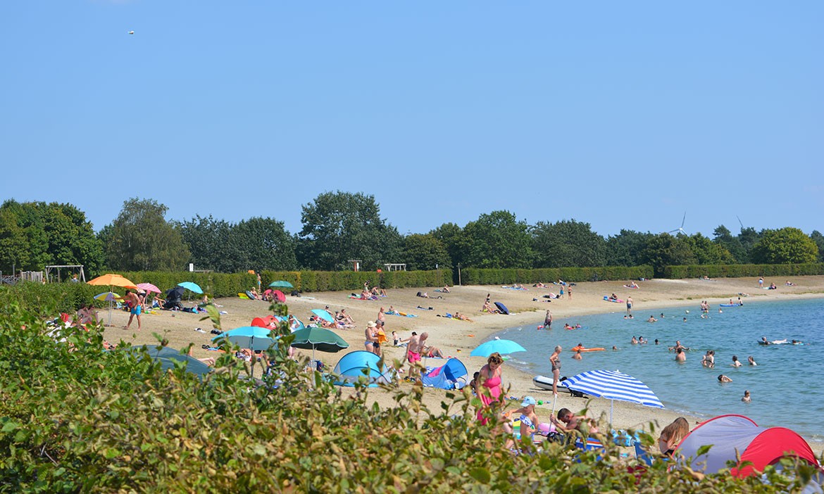 Sommerlicher Badestrand am Speichersee Geeste mit vielen Menschen am Sandufer und im flachen Wasser. Bunte Sonnenschirme, Handtücher und Badende prägen die Szene. Im Hintergrund schließen grüne Hecken und Bäume den Strandbereich ab.