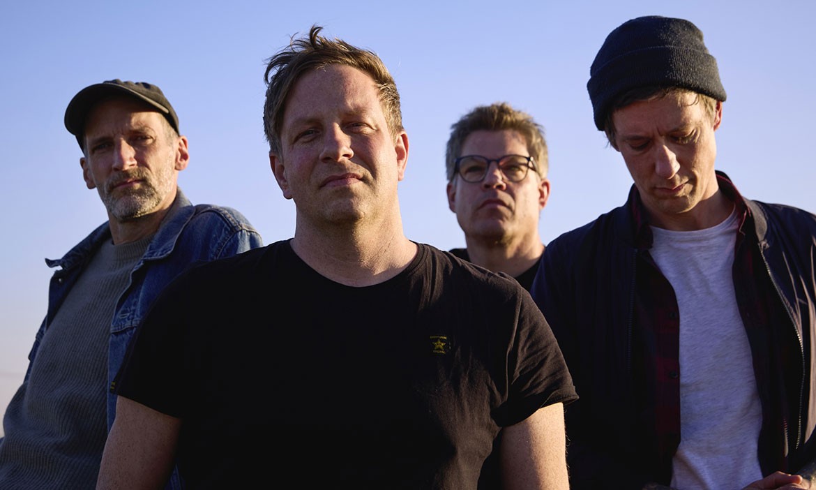 Die vier Mitglieder der Indie-Rock-Band Peals stehen unter freiem Himmel vor blauem Himmel. Die Musiker tragen dunkle, lässige Kleidung und blicken in die Kamera.