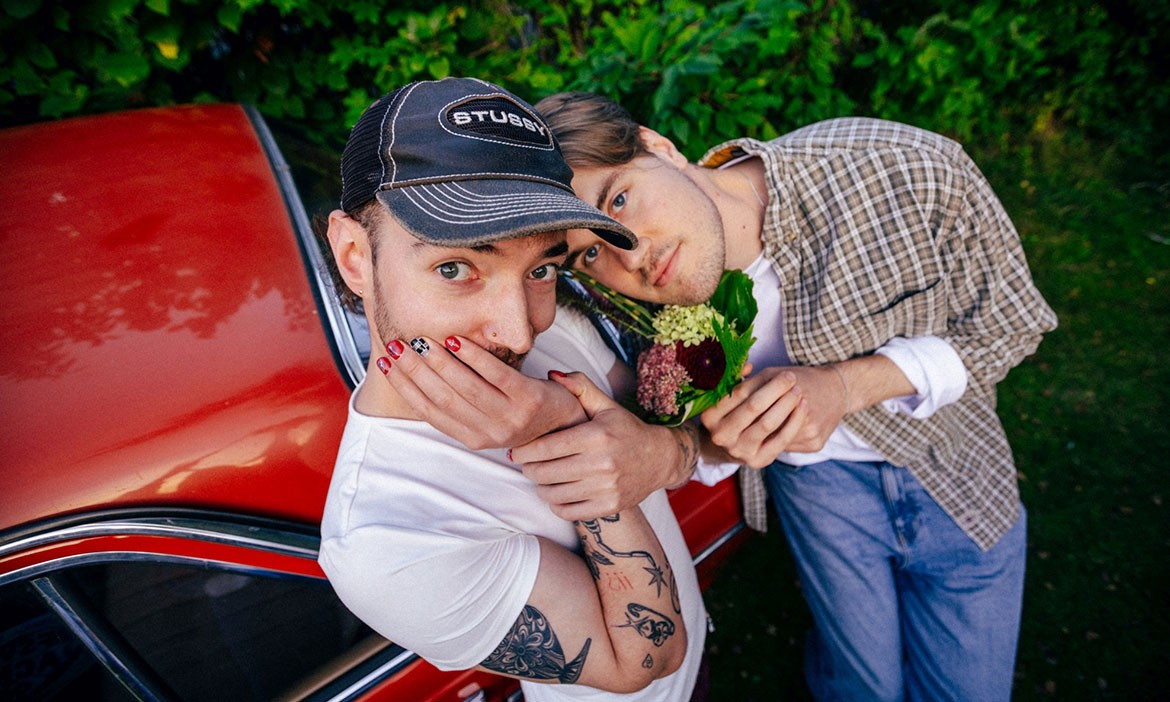 Die beiden Musiker der Indie-Pop-Band Raum27 posieren an einem roten Auto. Einer trägt eine schwarze Cap und zeigt lackierte Fingernägel, der andere hält einen kleinen Blumenstrauß in der Hand.