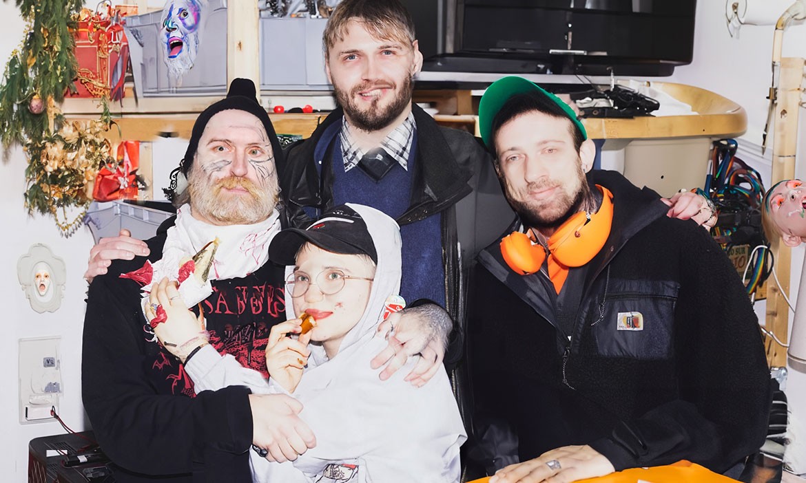 Die fünf Mitglieder der Punkband Team Scheisse posieren gemeinsam in einem ungewöhnlich dekorierten Raum. Die Gruppe trägt lässige Kleidung, Caps und auffällige Accessoires.