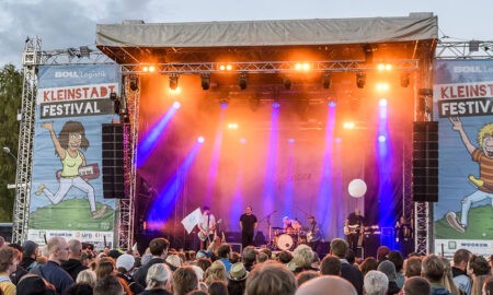 Große Open-Air-Bühne beim Kleinstadtfestival Meppen mit orangefarbenen und violetten Scheinwerfern. Eine Band spielt vor zahlreichen Festivalbesucher*innen, seitlich hängen große Kleinstadtfestival-Banner.