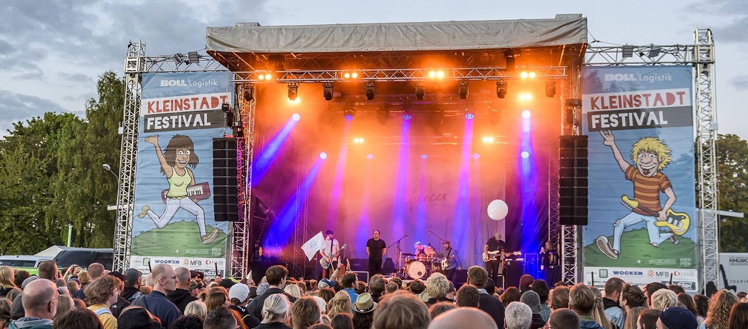 Große Open-Air-Bühne beim Kleinstadtfestival Meppen mit orangefarbenen und violetten Scheinwerfern. Eine Band spielt vor zahlreichen Festivalbesucher*innen, seitlich hängen große Kleinstadtfestival-Banner.