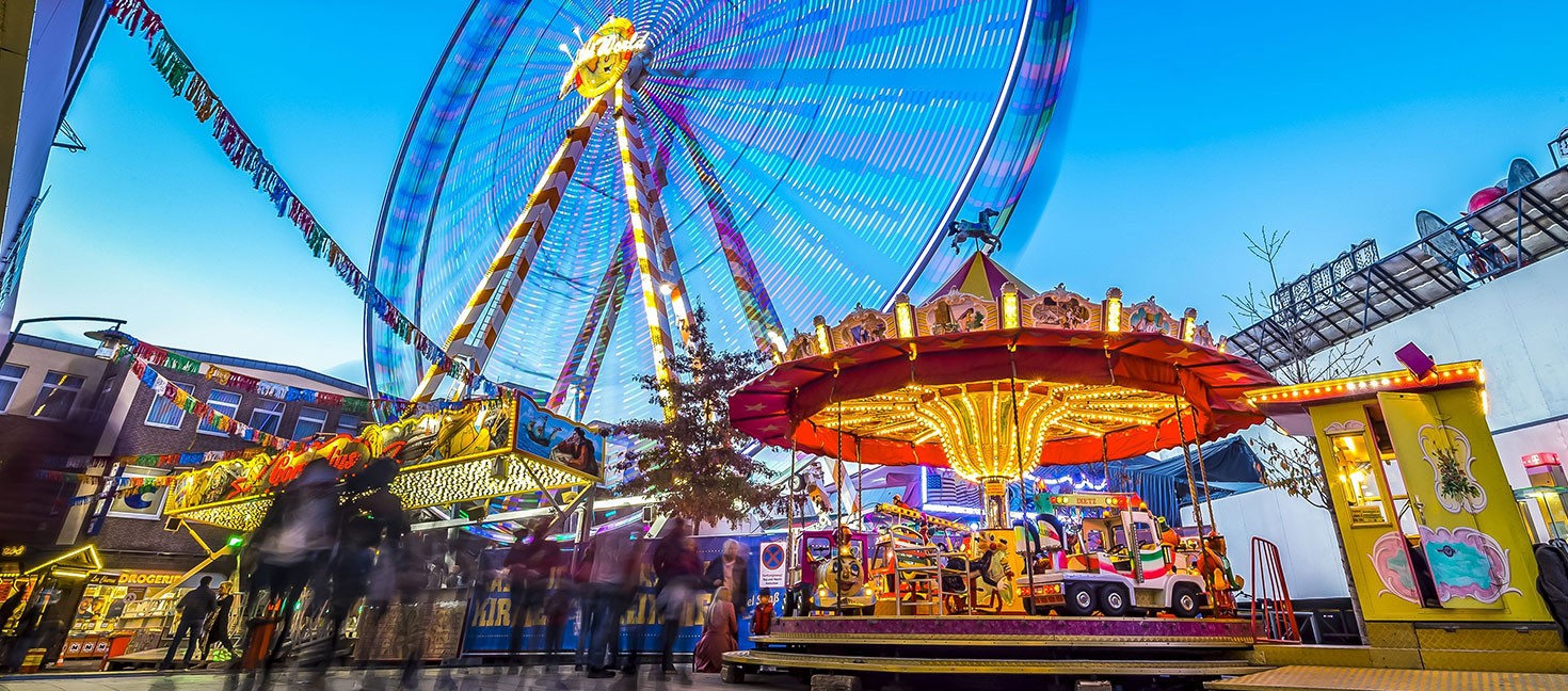 Bunt beleuchtetes Riesenrad und Kinderkarussell auf einem gut besuchten Jahrmarkt.