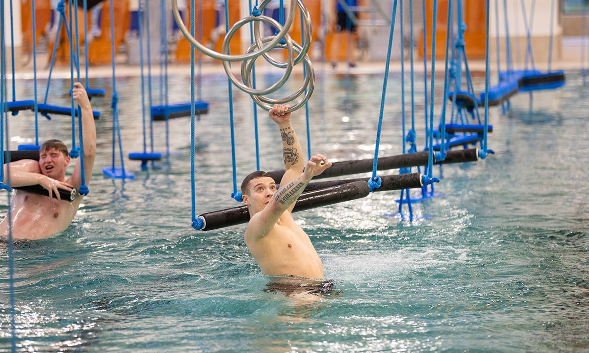 rueck_02.jpg	Ein Mann greift im Wasser nach hängenden Metallringen. Neben ihm versucht ein weiterer Mann, den Hindernisparcours über dem Becken zu überwinden.