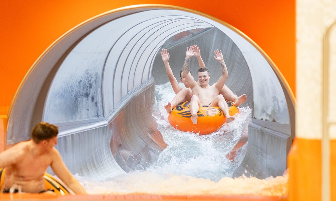Zwei Männer rutschen mit erhobenen Armen auf einem orangefarbenen Doppelreifen aus einer Wasserrutsche. Das Wasser spritzt um sie herum.