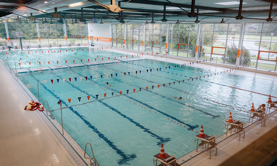Blick auf das Sportbecken im Nettebad Osnabrück mit abgetrennten Schwimmbahnen, Startblöcken und mehreren Schwimmenden. Große Fenster lassen Tageslicht in die Halle.